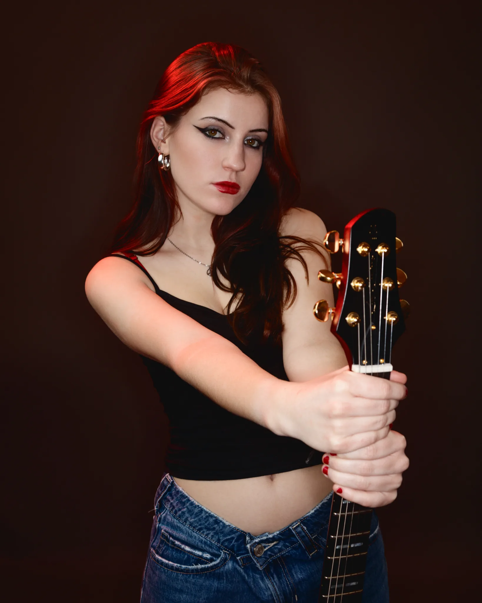 A woman with long red hair holds an acoustic guitar against a dark brown background. She wears a black crop top and blue jeans.